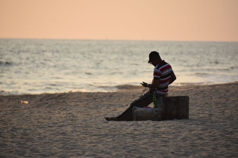 A Week Among The Wild Bonobos Of Congo A man sits on a log at a beach, facing the sea, wearing a striped shirt and a cap, looking at a cell phone in his hand.